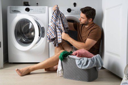 Glücklicher Mann, der in der Waschküche neben der Waschmaschine Wäsche sortiert. Mann sitzt auf dem Boden eines Hauses in der Nähe einer Waschmaschine mit schmutziger Kleidung. ein Mann wäscht seine Wäsche zu Hause. ein Mann mit schmutziger Kleidung
