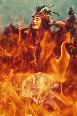 Photo for Beautiful shamanic girl playing on shaman frame drum in the nature. Fire background - Royalty Free Image