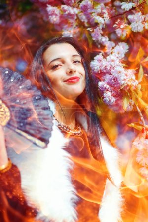 Photo for Against a backdrop of blooming flowers, a woman stands confidently. She resembles an infernal fashion show attendee, an evil fire sorceress. Flames envelop her, accentuating her fiery presence. - Royalty Free Image