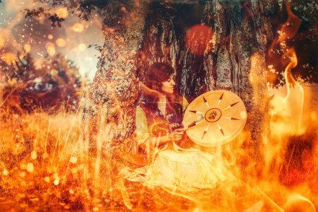 Foto de Hermosa chica chamánica jugando en tambor marco chamán en la naturaleza. Fondo de fuego - Imagen libre de derechos
