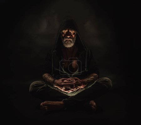 Photo for Meditating man in a hood sits on the ground, man on the hands of the tattoo, atelier photos, black background - Royalty Free Image