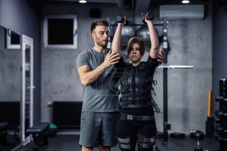 Female and male confident athletes train indoors at the gym. Woman is dressed in EMS technology in a gym and lifts dumbbells over her head while her trainer helps her perform a new exercise concept