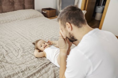 Un père stupide joue à jeter un coup d'oeil avec sa fille à la maison pendant qu'elle se couche sur le lit.