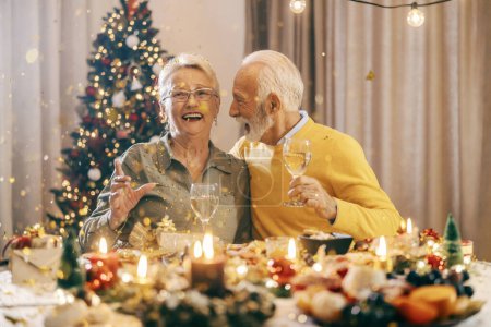 A festive excited grandparents celebrating christmas and new year's eve with confetti and hugging at home at dining table.