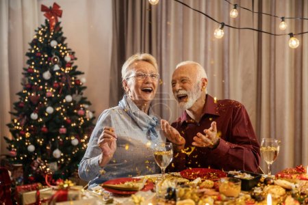 Excited senior couple is celebrating christmas and new year's eve at midnight with confetti at home at dining table.