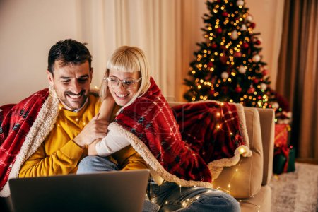 A happy couple is sitting at cozy home on christmas and new year's eve and smiling at the laptop.