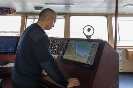 Officer on watch with electronic chart on the navigational bridge. Caucasian man in blue uniform sweater using ECDIS on the bridge of cargo ship.