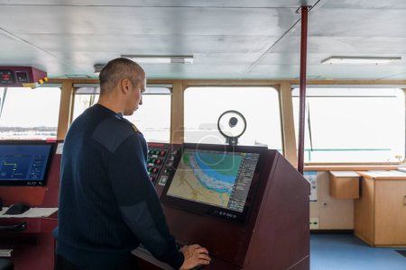 Officer on watch with electronic chart on the navigational bridge. Caucasian man in blue uniform sweater using ECDIS on the bridge of cargo ship.