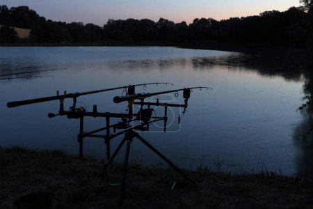 Foto de La puesta de sol sobre el horizonte y la pesca junto al estanque. Ponte con cañas de pescar. La superficie del lago. - Imagen libre de derechos