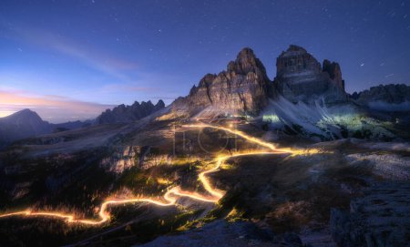 Foto de Senderos ligeros para coches en carretera de montaña y rocas altas por la noche en otoño en Tre cime, Dolomitas, Italia. Paisaje colorido con senderos de luz borrosa, colinas, picos de montaña, cielo con estrellas en otoño al atardecer - Imagen libre de derechos