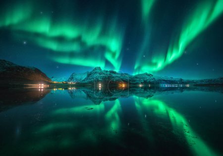 Northern lights over the snowy mountains, sea, reflection in water at night in Lofoten, Norway. Aurora borealis and snow covered rocks. Winter landscape with polar lights, city lights, sky with stars