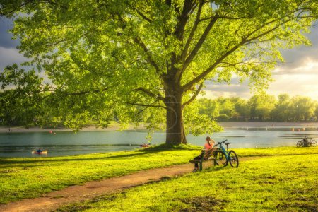 Photo for Woman sitting on bench and mountain bike, green trees and grass, lake at sunset in spring. Colorful landscape with resting girl, bicycle, river in park in summer. Sport and travel. Biking. Nature - Royalty Free Image