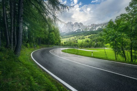 Téléchargez les photos : Route en forêt verte en été pluvieux. Dolomites, Italie. Belle route de montagne, teresse, herbe, hauts rochers, ciel bleu avec nuages. Paysage avec autoroute vide à travers le bois au printemps. Voyages - en image libre de droit