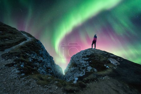 Foto de Luces boreales y mujer joven en el pico de la montaña por la noche. Aurora boreal verde y violeta y silueta de chica solitaria en sendero de montaña. Paisaje con luces polares. Cielo estrellado con aurora brillante - Imagen libre de derechos
