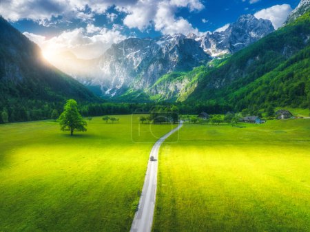 Aerial view of road, alpine mountains, green meadows, trees in summer at sunset. Top view of rural road. Landscape with road, car, rocks, field, grass, sky, clouds in spring. Logar valley, Slovenia