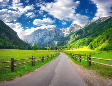 Rural road in alpine mountains, wooden fence, green meadows, trees in summer in Logar valley, Slovenia. Country road. Colorful landscape with road, rocks, field, grass, blue sky with clouds at sunset