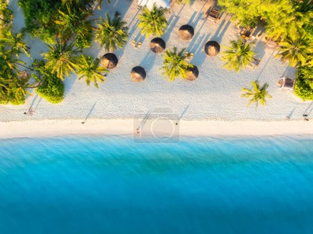 Luftaufnahme von grünen Palmen, Sonnenschirmen am Sandstrand des Indischen Ozeans bei Sonnenuntergang. Sommerurlaub in Kendwa, Insel Sansibar. Tropische Landschaft mit Palmen, weißem Sand, klarem blauen Meer. Ansicht von oben