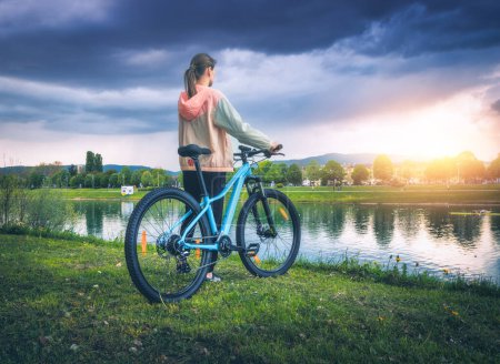 Frau mit Mountainbike am See bei bedecktem Sonnenuntergang im Frühling. Bunte Landschaft mit sportlichem Mädchen, Fahrrad, Flussküste, grünem Gras, bewölktem Himmel im Sommer im Park. Sport und Reisen. Radfahren