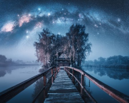 Photo for Wooden walkway and house surrounded by trees, Milky Way galaxy in the night sky in Ukraine. Landscape with calm river reflecting the trees and house in fog, starry sky with glowing Milky Way arch - Royalty Free Image