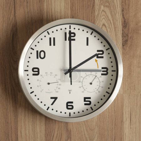 A clock, the gray hand points to three o'clock, the black one to two o'clock. The orange arrow indicates the direction of movement of the hands. Symbol of the time change. Standard time.