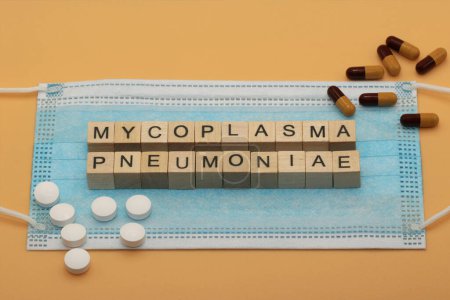Photo for Mycoplasma pneumoniae. It is a human pathogen that causes the disease mycoplasma pneumonia. The words are laid out with wooden cubes on a surgical face mask. There are various pills lying around. - Royalty Free Image