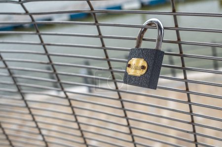 Single grey padlock on a wire mesh fence with gold lable with out of focus background.