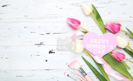 Photo for Flowers of tulips, card in shape of heart with text Happy Women's Day, pink makeup brushes and ribbon on white wooden background - Royalty Free Image