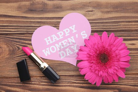 Téléchargez les photos : Gerbera fleur, rouge à lèvres et carte en forme de coeur avec texte Happy Women's Day sur fond brun en bois - en image libre de droit