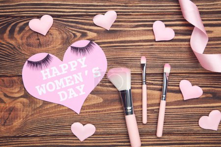 Photo for Card in shape of heart with text Happy Women's Day, pink ribbon and hearts, set makeup brushes, eyelashes on brown wooden background - Royalty Free Image