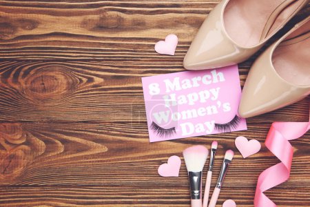 Foto de Tarjeta con texto 8 Marzo Feliz Día de la Mujer, cinta rosa y corazones, set pinceles de maquillaje y pestañas, par de zapatos de tacón alto beige sobre fondo de madera marrón - Imagen libre de derechos