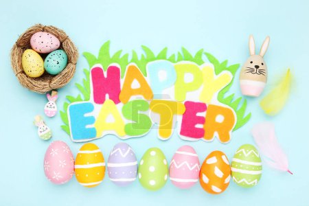 Téléchargez les photos : Œufs colorés et inscription colorée Joyeux Pâques, lapin et plumes sur fond bleu - en image libre de droit