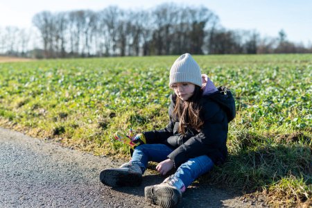 Little girl sitting on the road in the field and playing with grass