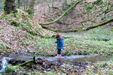 Kleines Mädchen in Regenmantel und Gummistiefeln steht in einem kleinen Bach im Wald.