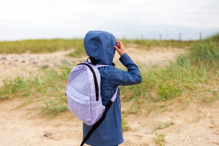 Back view of a little girl in a blue raincoat with a backpack on the beach