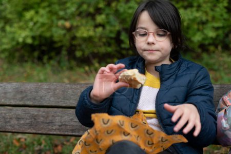 Little girl sitting on a bench in the park and eating a sandwich