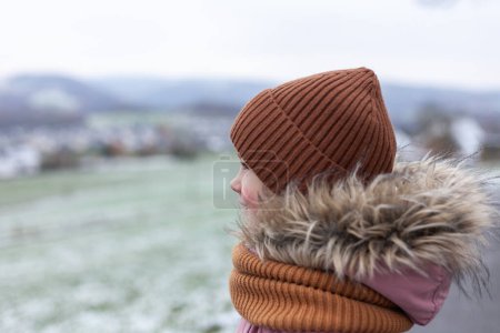 Ein kleines Mädchen mit warmer Mütze und Schal steht auf dem Hintergrund einer Winterlandschaft.