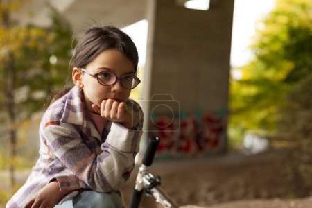 A tired little girl in a plaid shirt and glasses leaned her head on her hand while sitting under a bridge next to a scooter