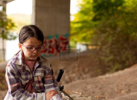 Nettes kleines Mädchen in kariertem Hemd und Brille sitzt traurig unter einer Brücke