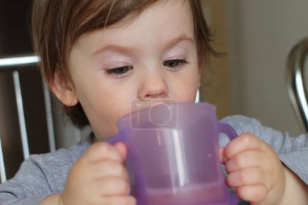 Un niño pequeño bebe de una taza con dos manos. Aprender a beber de una taza de forma independiente. Hitos del desarrollo y la alegría de la vida familiar cotidiana
