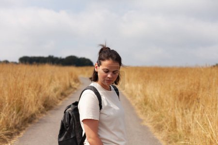 Junge Frau mit Rucksack auf einer Landstraße, umgeben von der Schönheit eines goldenen Weizenfeldes und umarmt das Gefühl von Abenteuer und Freiheit, das mit der Erkundung der Landschaft einhergeht