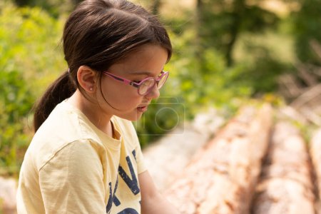 Ein kleines hübsches Mädchen mit Brille und Pferdeschwanz sitzt lächelnd auf den abgesägten Stämmen einer Kiefer. BLUR natürlicher Hintergrund