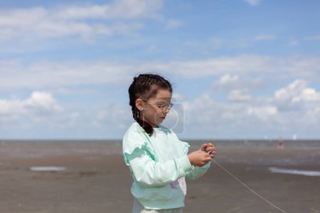 Kleines Mädchen in Kleidern steht mit Drachenschnur am Strand