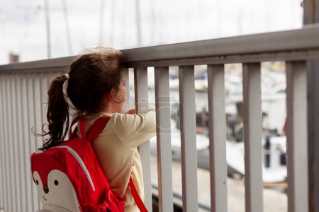 A little girl with a backpack stands at the railing looking at the ships in the harbor