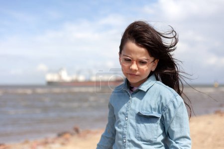 Ein kleines Mädchen mit Brille steht am Strand. Großes Schiff für den Gütertransport im Hintergrund