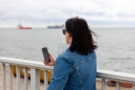 A girl in a dark denim jacket talks on a video call on the North Sea embankment