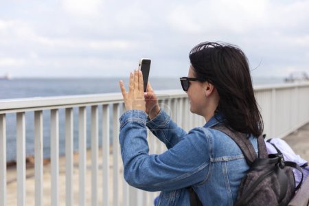 Ein nettes dunkelhaariges Mädchen in Jeansjacke macht Fotos mit ihrem Handy, während sie an der Küste steht