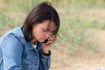 Ein schönes dunkelhaariges Mädchen in Jeansjacke telefoniert traurig am Strand in der Nähe des Meeres