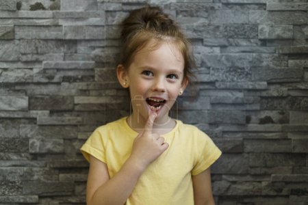 Photo for Little girl in a yellow T-shirt on a brick wall background points to a lost tooth - Royalty Free Image