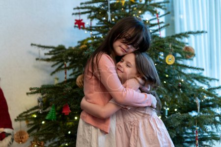 Niña y su hermana se abrazan cerca del árbol de Navidad.
