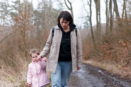 A young girl with a backpack walks hand in hand with her little girl along a dirty forest winter road